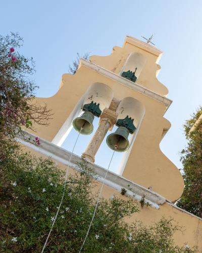 A low-angle shot of a yellow church bell tower with three bells, set against a blue sky and surrounded by green vegetation with white and pink flowers.