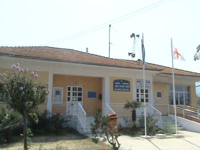 A single-story yellow building with a tiled roof, identified by Greek signage as a medical facility (specifically "ΔΗΜΟΤΙΚΟ ΙΑΤΡΕΙΟ Ν. ΠΟΡΩΝ" - Municipal Clinic of Neoi Poroi), with a Greek flag and a red cross flag flying outside.