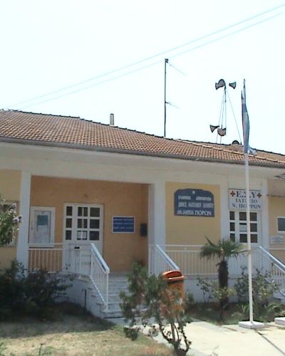 A single-story yellow building with a tiled roof, identified by Greek signage as a medical facility (specifically "ΔΗΜΟΤΙΚΟ ΙΑΤΡΕΙΟ Ν. ΠΟΡΩΝ" - Municipal Clinic of Neoi Poroi), with a Greek flag and a red cross flag flying outside.