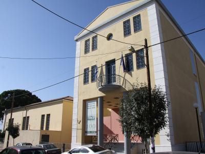 A yellow neoclassical building with white trim, Greek flags, and a banner reading "ΑΡΧΑΙΟΛΟΓΙΚΟ ΜΟΥΣΕΙΟ ΒΟΛΟΥ" (Archaeological Museum of Volos) and "100 ΧΡΟΝΙΑ" (100 Years).
