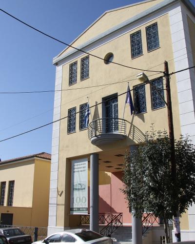 A yellow neoclassical building with white trim, Greek flags, and a banner reading "ΑΡΧΑΙΟΛΟΓΙΚΟ ΜΟΥΣΕΙΟ ΒΟΛΟΥ" (Archaeological Museum of Volos) and "100 ΧΡΟΝΙΑ" (100 Years).