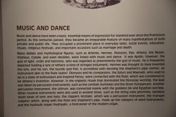 A close-up photograph of an informational museum placard with the heading "MUSIC AND DANCE" and text describing ancient Greek musical instruments and mythology.