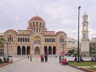 A large Byzantine-style cathedral with a central dome and red-tiled roofing, situated in a paved public square with a separate tall bell tower to the right and people walking in the foreground.