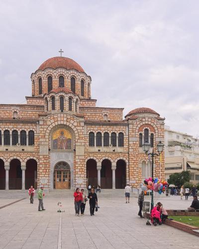 A large Byzantine-style cathedral with a central dome and red-tiled roofing, situated in a paved public square with a separate tall bell tower to the right and people walking in the foreground.