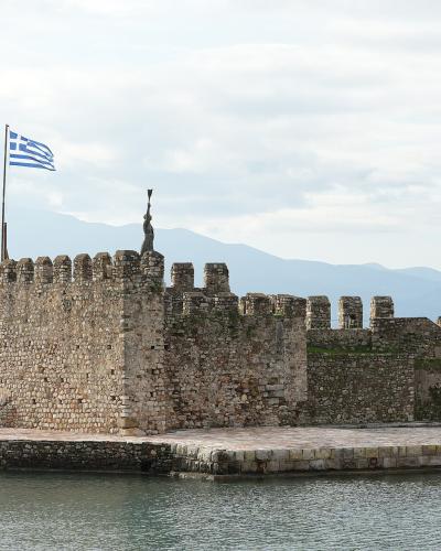 A stone fortress wall extending into the water with a Greek flag flying from a pole and a statue on top, alongside a small boat moored at the quay.