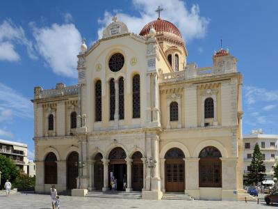 A large, cream-colored cathedral with a central red dome, arched windows, and a stone facade under a blue sky, with people walking in the plaza in front.