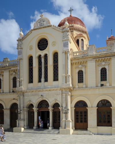 A large, cream-colored cathedral with a central red dome, arched windows, and a stone facade under a blue sky, with people walking in the plaza in front.