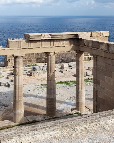 The image shows the ancient stone ruins of a Greek temple portico with four fluted columns and a section of entablature, situated on a rocky cliff overlooking the blue Aegean Sea.
