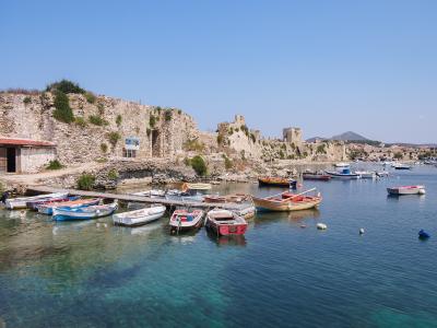 A view of a historic stone fortress wall running along a harbor filled with small fishing boats and yachts under a clear blue sky.