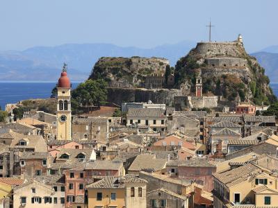A panoramic view of the Old Town of Corfu with its distinctive terracotta-roofed buildings and the Old Fortress (Palaio Frourio) situated on a rocky promontory overlooking the sea, with the Church of Saint Mark's bell tower visible in the foreground.