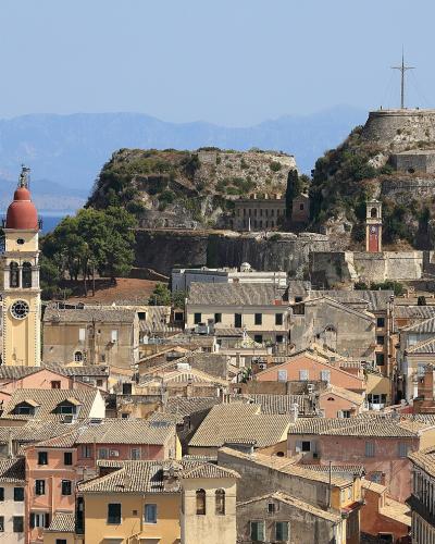 A panoramic view of the Old Town of Corfu with its distinctive terracotta-roofed buildings and the Old Fortress (Palaio Frourio) situated on a rocky promontory overlooking the sea, with the Church of Saint Mark's bell tower visible in the foreground.