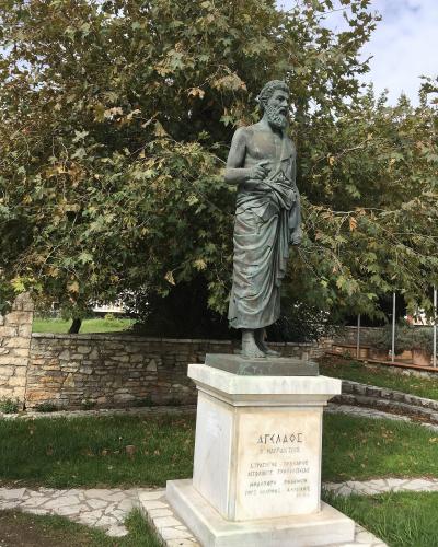 A bronze statue of a bearded man in classical robes standing on a marble pedestal with Greek inscriptions, set outdoors against a backdrop of trees and a stone wall.