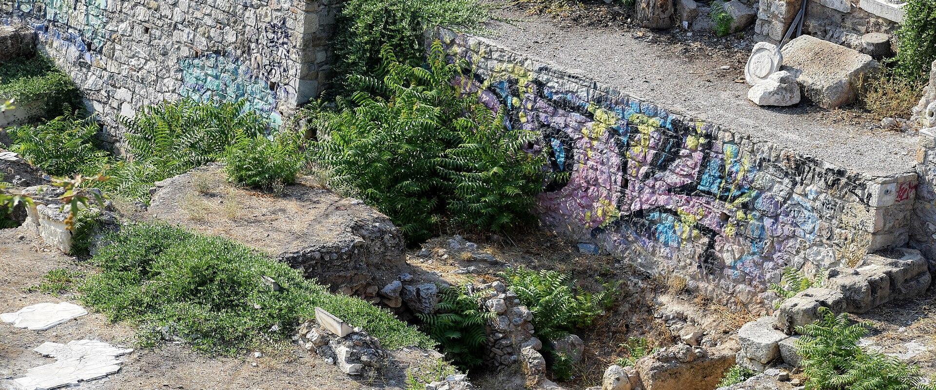 An archaeological excavation site featuring ancient stone foundations and walls covered in colorful graffiti, with overgrown vegetation.