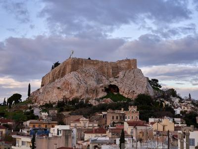 A view of the Acropolis of Athens rising above the rooftops of a residential neighborhood, featuring the Propylaea entrance structure and the cave of Aglauros on the rock face.