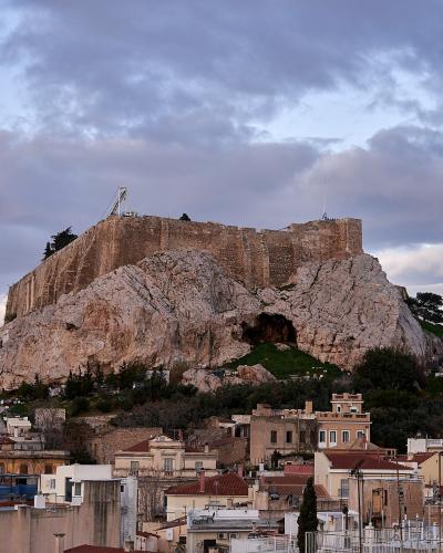 A view of the Acropolis of Athens rising above the rooftops of a residential neighborhood, featuring the Propylaea entrance structure and the cave of Aglauros on the rock face.