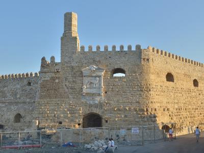 A stone Venetian fortress with crenellated walls, a central tower, and a prominent sculpted relief of a winged lion on the facade, situated by a harbor with a boat visible in the background.