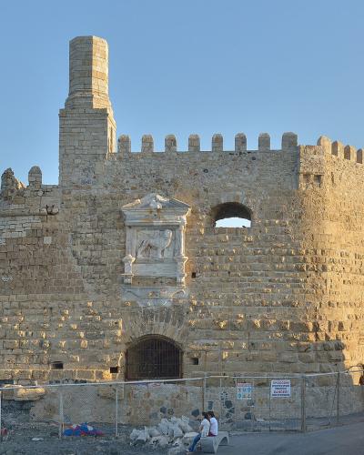 A stone Venetian fortress with crenellated walls, a central tower, and a prominent sculpted relief of a winged lion on the facade, situated by a harbor with a boat visible in the background.