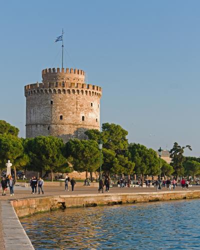 A close-up view of the White Tower of Thessaloniki, a cylindrical stone fortress situated on the waterfront promenade with a Greek flag flying from its top, surrounded by green trees and pedestrians.