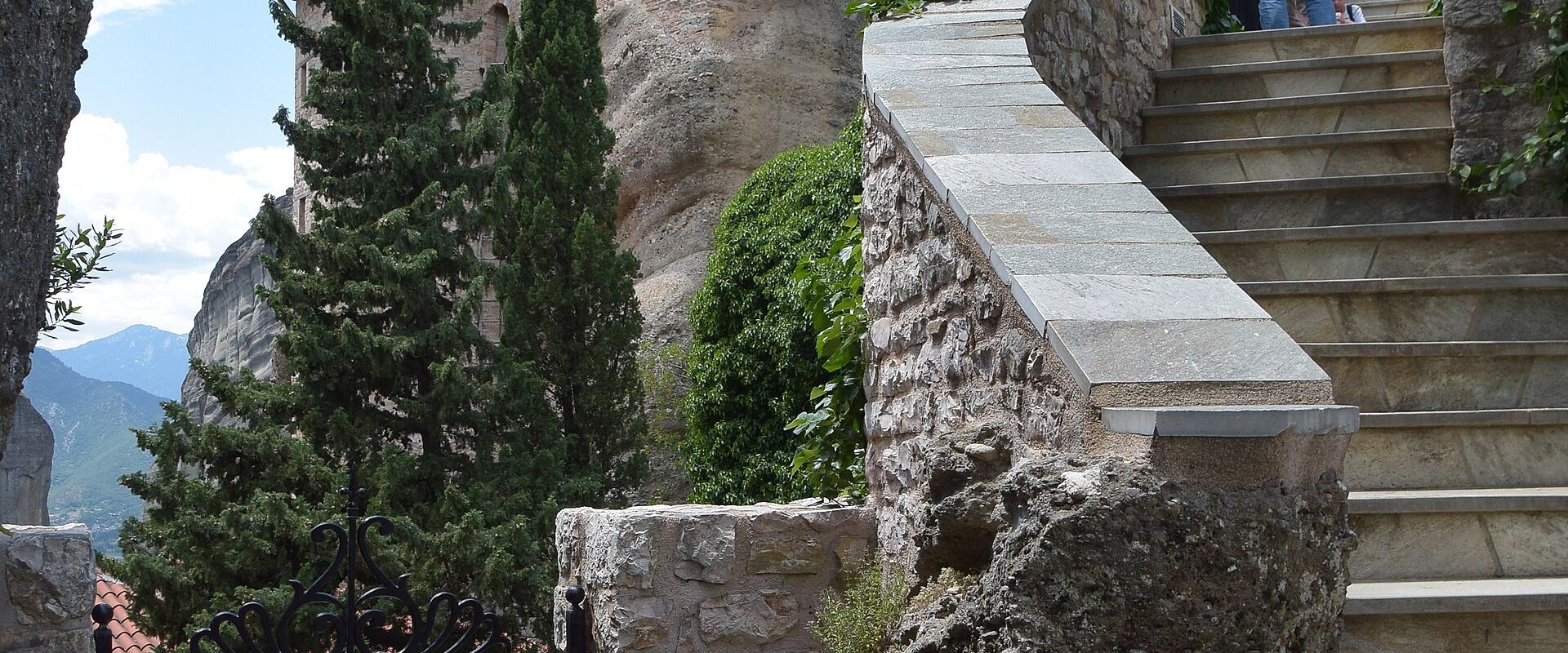 A stone monastery building built into a rock face with a wooden balcony, accessed by a stone staircase, with a black metal gate in the foreground bearing a sign with Greek text.
