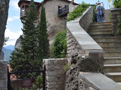A stone monastery building built into a rock face with a wooden balcony, accessed by a stone staircase, with a black metal gate in the foreground bearing a sign with Greek text.