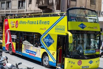 A yellow and blue double-decker sightseeing bus branded "Gray Line by Athens Open Tour" with text reading "Athens-Piraeus & Riviera" and "TO ACROPOLIS" on the front, parked on a city street.