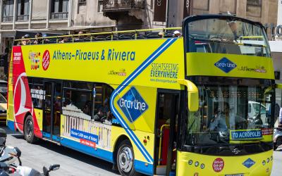 A yellow and blue double-decker sightseeing bus branded "Gray Line by Athens Open Tour" with text reading "Athens-Piraeus & Riviera" and "TO ACROPOLIS" on the front, parked on a city street.