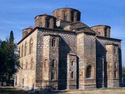 The image shows a large, historic Byzantine-style church constructed of stone and brick, featuring a central dome flanked by smaller semi-domes, set against a clear blue sky with trees visible to the left.