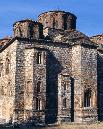 The image shows a large, historic Byzantine-style church constructed of stone and brick, featuring a central dome flanked by smaller semi-domes, set against a clear blue sky with trees visible to the left.
