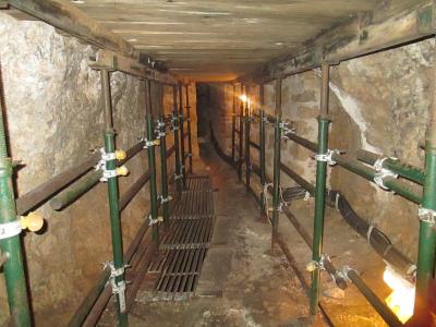 A view inside a narrow, rough-hewn stone tunnel with wooden ceiling beams and modern green metal shoring supports lining the walls.
