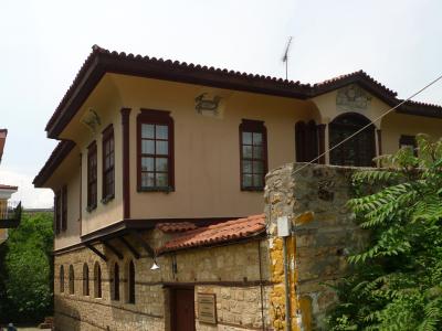 A two-story building with a stone ground floor and a stucco upper floor featuring dark wood window frames and decorative elements, with a small plaque mounted on the stone wall near the entrance.