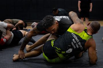 The image shows an indoor martial arts training session with two men grappling on mats, one wearing a shirt that reads "KYDONAS SPORTS MUAY THAI CHANIA".