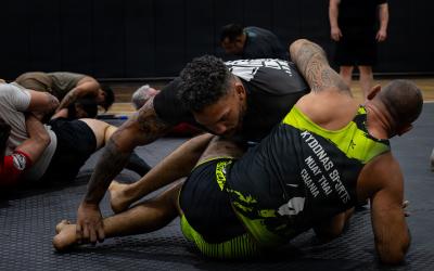 The image shows an indoor martial arts training session with two men grappling on mats, one wearing a shirt that reads "KYDONAS SPORTS MUAY THAI CHANIA".