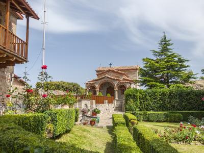 A view of a stone monastery building with a terracotta-tiled roof and arched portico, set within a manicured garden featuring hedges, rose bushes, and a wooden balcony structure on the left.