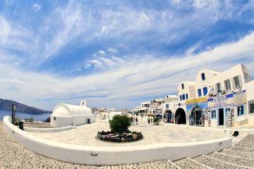 A wide-angle view of a paved town square in a Greek island setting, featuring a white domed church on the left, a cobblestone foreground, and a row of white buildings with blue accents on the right, including a shop with a sign reading "DROSSOS CAR &