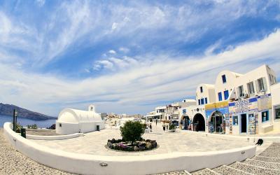 A wide-angle view of a paved town square in a Greek island setting, featuring a white domed church on the left, a cobblestone foreground, and a row of white buildings with blue accents on the right, including a shop with a sign reading "DROSSOS CAR &