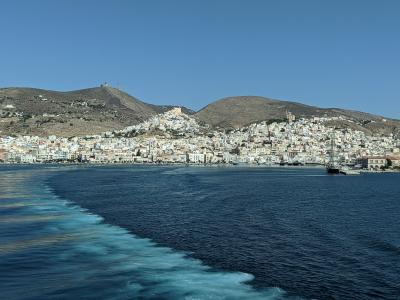 A coastal town with white buildings climbing a hillside, viewed from the wake of a moving boat on the water.
