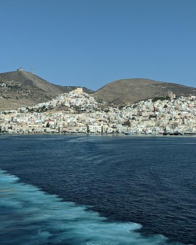 A coastal town with white buildings climbing a hillside, viewed from the wake of a moving boat on the water.