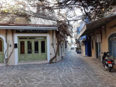 A narrow, cobblestone pedestrian street in a town, featuring a cafe named "KANTOUNI" on the left and a scooter parked on the right, with vines overhead.
