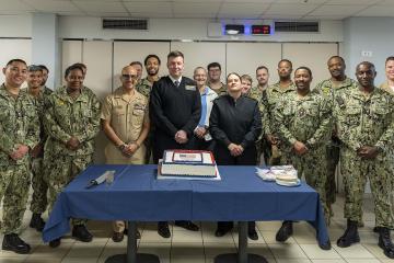 A group of U.S. Navy personnel in various uniforms posing indoors behind a table with a commemorative cake.