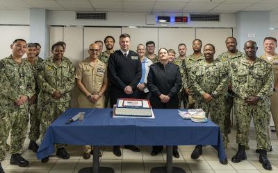 A group of U.S. Navy personnel in various uniforms posing indoors behind a table with a commemorative cake.