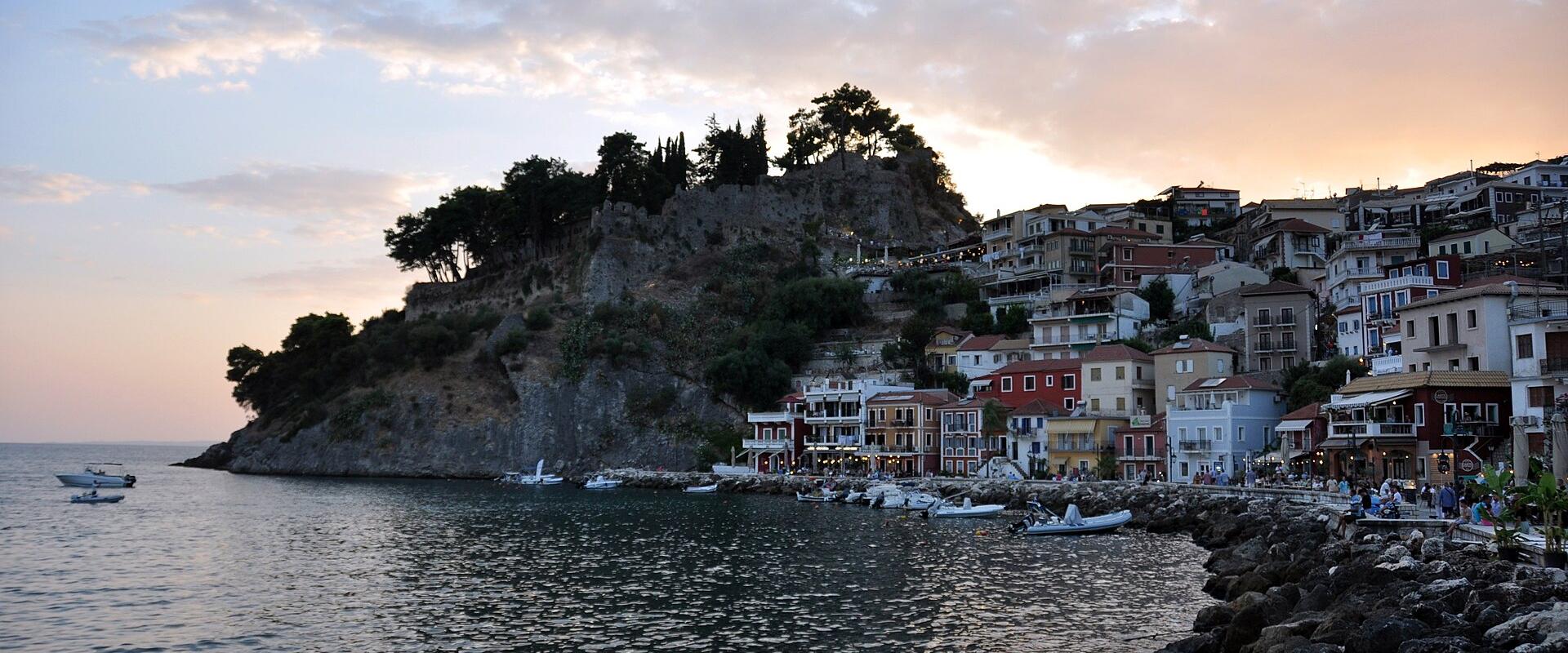 The image shows a coastal town at dusk, featuring a prominent rocky islet or headland topped with trees and a stone structure (likely a castle or fortress) on the left, while the right side shows a shoreline with multi-story buildings, a promenade, a