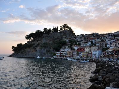 The image shows a coastal town at dusk, featuring a prominent rocky islet or headland topped with trees and a stone structure (likely a castle or fortress) on the left, while the right side shows a shoreline with multi-story buildings, a promenade, a