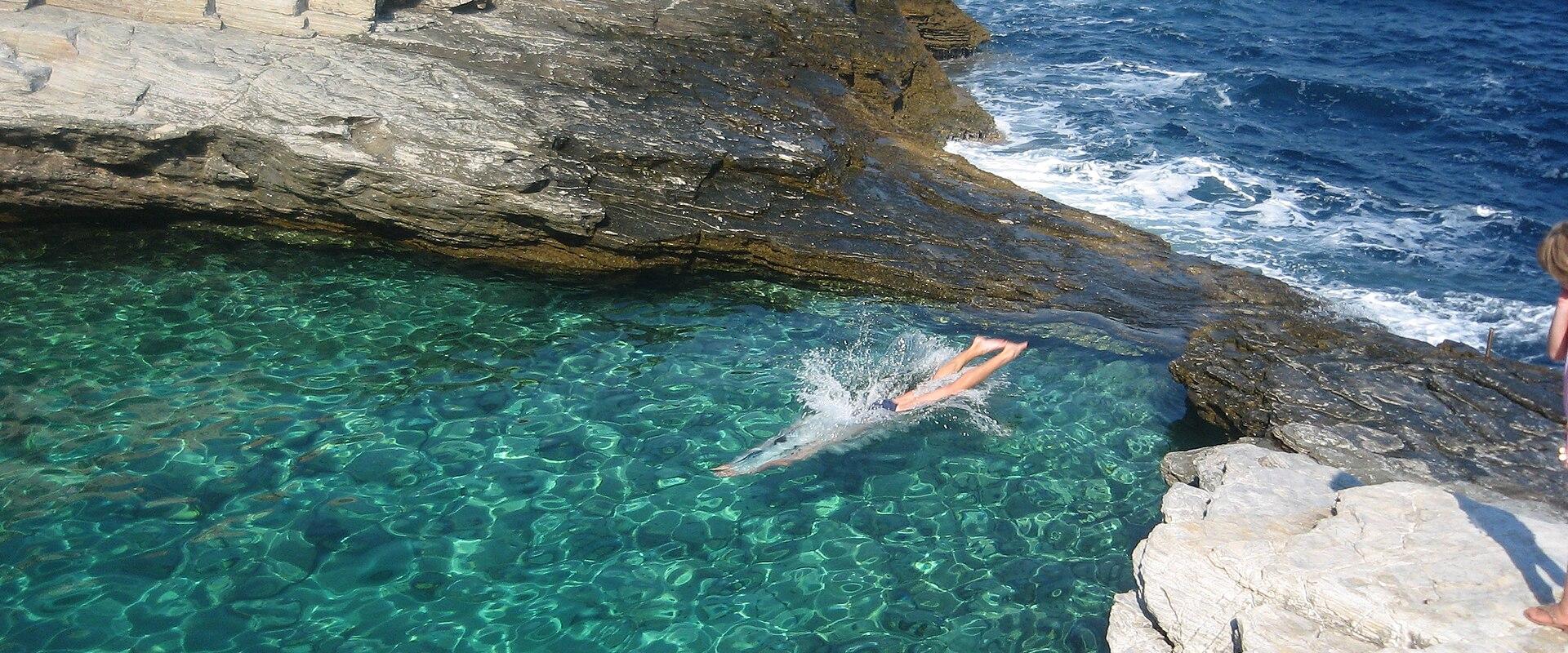 A person is diving into a natural rock pool with clear turquoise water, surrounded by layered limestone cliffs, with the open blue sea visible in the background.