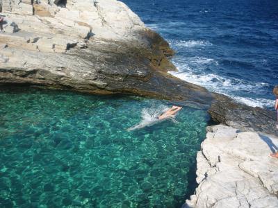 A person is diving into a natural rock pool with clear turquoise water, surrounded by layered limestone cliffs, with the open blue sea visible in the background.