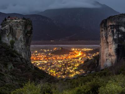 A night view of the town of Kalabaka illuminated between two massive sandstone rock pillars, with a monastery visible on the summit of the left pillar.