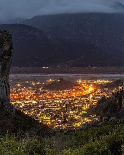 A night view of the town of Kalabaka illuminated between two massive sandstone rock pillars, with a monastery visible on the summit of the left pillar.