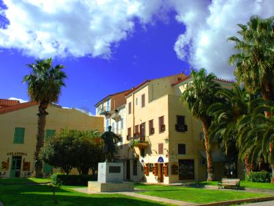 The image shows a public square with a central statue of a man on a pedestal, surrounded by green grass, palm trees, and a building with a "GELATERIA" sign.