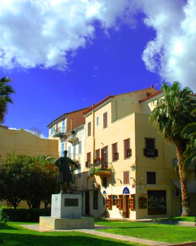 The image shows a public square with a central statue of a man on a pedestal, surrounded by green grass, palm trees, and a building with a "GELATERIA" sign.