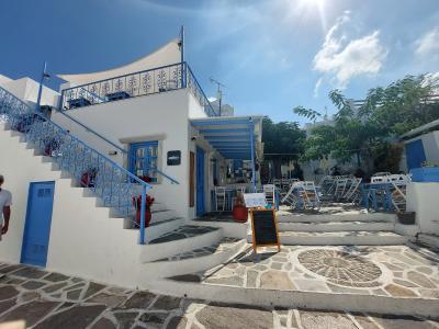 A white-washed Cycladic building with blue trim, featuring an external staircase, a small cafe or taverna seating area with white chairs, and a stone-paved square in the foreground.