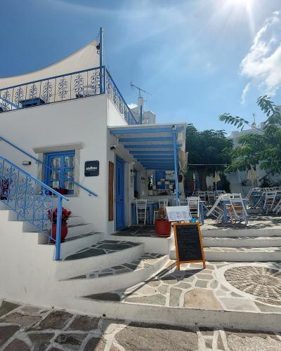 A white-washed Cycladic building with blue trim, featuring an external staircase, a small cafe or taverna seating area with white chairs, and a stone-paved square in the foreground.